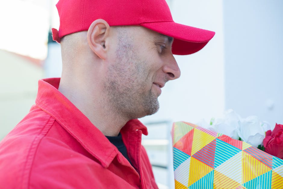 A man in a red cap and matching red jacket holding a brightly wrapped floral gift with a geometric multicolored pattern, containing white and red flowers, during daylight. The bouquet appears fresh wi