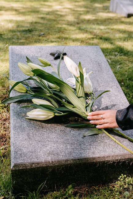 A hand holding a bouquet of fresh, predominantly white lilies with elongated petals and green foliage, being placed on a flat, polished grey granite gravestone at a cemetery. The arrangement includes 