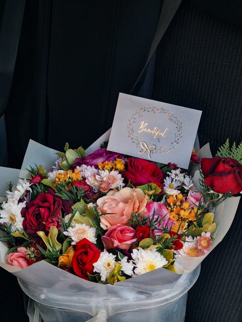 A vibrant floral arrangement featuring a mix of roses, peonies, daisies, and other fresh flowers in shades of red, pink, white, yellow, and purple, arranged in a round bouquet with green foliage accen