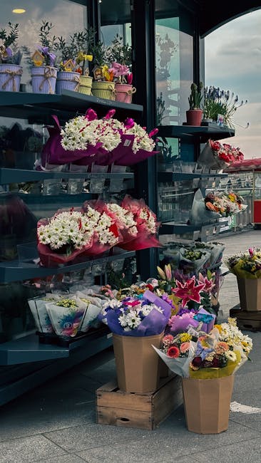 A display of various fresh floral arrangements on outdoor metal shelves and a wooden crate, featuring bouquets of white, pink, purple, and yellow flowers wrapped in colorful paper and tissue. The arra