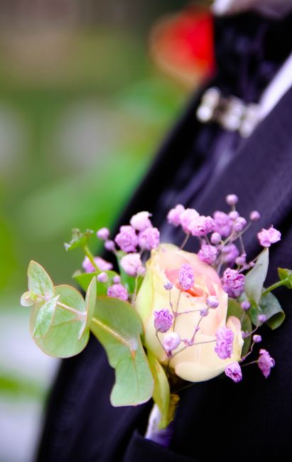 A boutonniere featuring a single pale peach rose accompanied by clusters of delicate purple baby's breath and green eucalyptus leaves, arranged with fresh, vibrant petals and sturdy stems. The floral