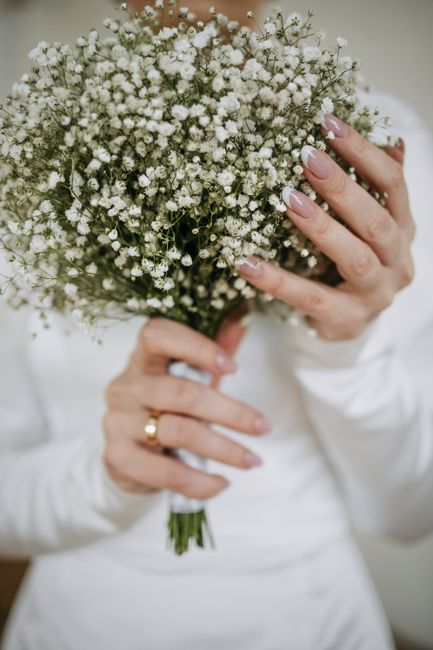 A person holding a round bouquet of delicate white baby's breath flowers with lush green stems, wrapped in white tissue paper. The bouquet showcases a soft, airy texture, with tiny white blooms densel